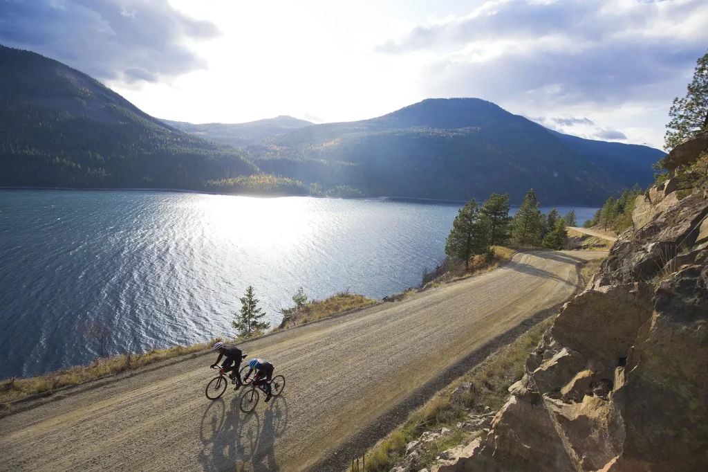 Two people biking down a road by a lake and hills