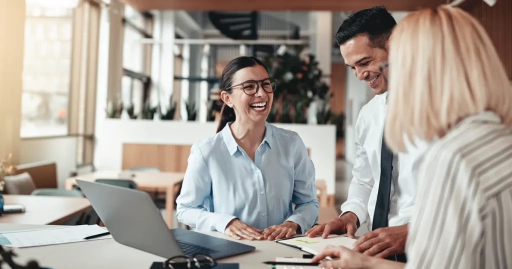 A smiling professional meeting with colleagues around a table with a laptop
