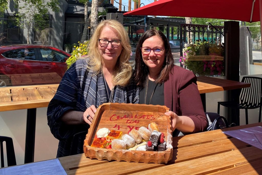 Susyn Wagner and Karen Routledge smiling at an outdoor table, holding a wooden platter with dessert and the message ‘Congrats on 4 Years’ written in sauce.